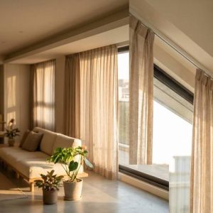 Bright living room with beige curtains, large angled window, sunlit sofa, and potted plants, creating a warm atmosphere.