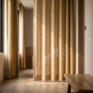 Sunlit hallway with tall beige curtains along windows, wooden floor, and a small bench on the right end of the corridor.