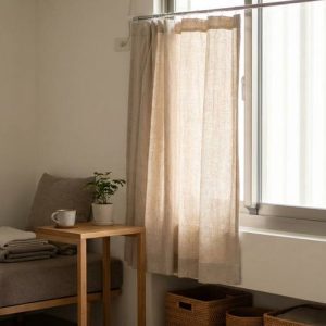 Small bedroom corner with a single bed, beige linens, a wooden bedside table, potted plant, and a window with sheer curtains.