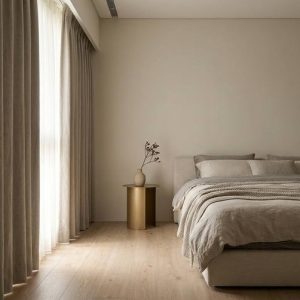 Minimalist bedroom with a low bed, neutral linens, and a small round side table with a vase and dried branches; floor-to-ceiling beige curtains on the left behind the bed.