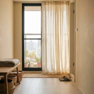 Bright apartment entryway with a bench, folded towels, woven baskets, and a balcony door with sheer curtains.