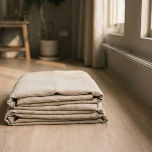 Folded beige blankets stacked neatly on a wooden floor by a sunlit living room window
