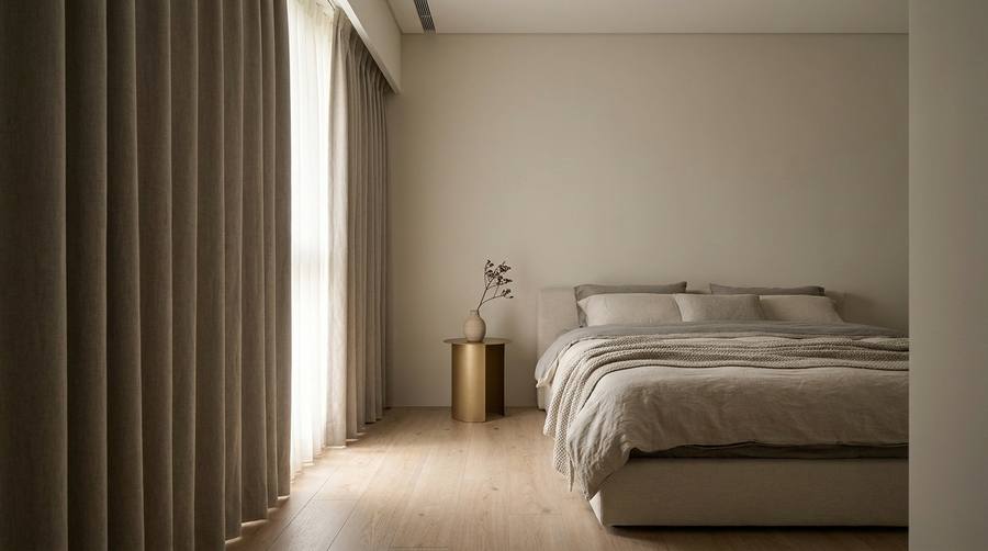 Minimalist bedroom with a low bed, neutral linens, and a small round side table with a vase and dried branches; floor-to-ceiling beige curtains on the left behind the bed.