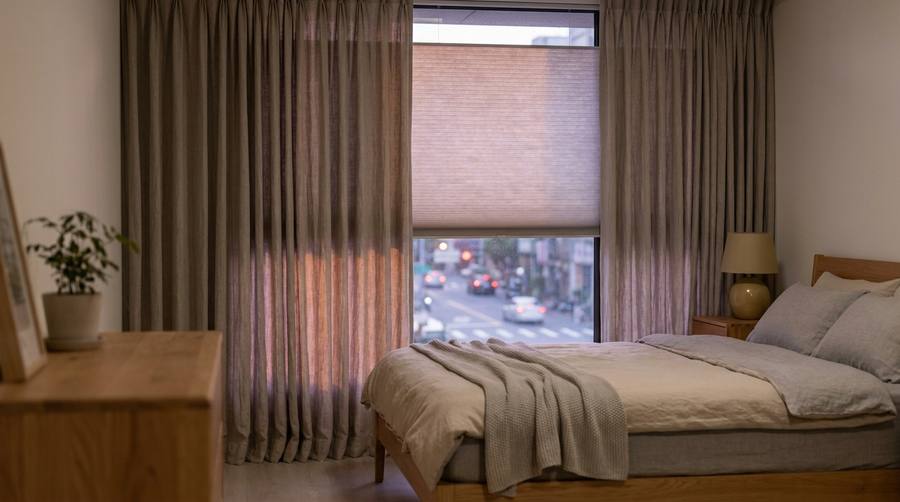 Cozy bedroom with a wooden bed, beige bedding, and a large window framed by sheer curtains. A dresser with a plant and a bedside lamp sit nearby.