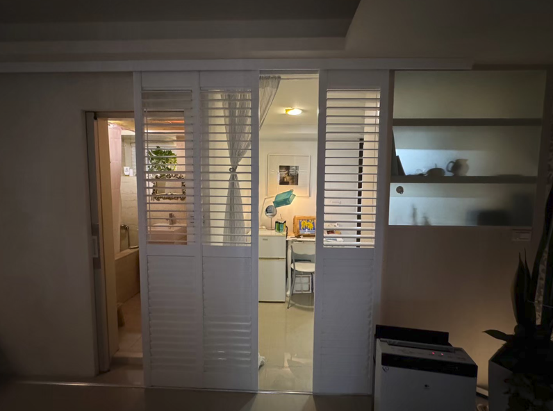 臥室的窗戶近景。窗戶安裝了雙層窗簾：內層是白色或米白色的柔紗簾/紗簾，外層是淺灰色或淺米色的落地布窗簾，兩者都呈現垂直的波浪褶皺。左側有一張深色皮質單椅