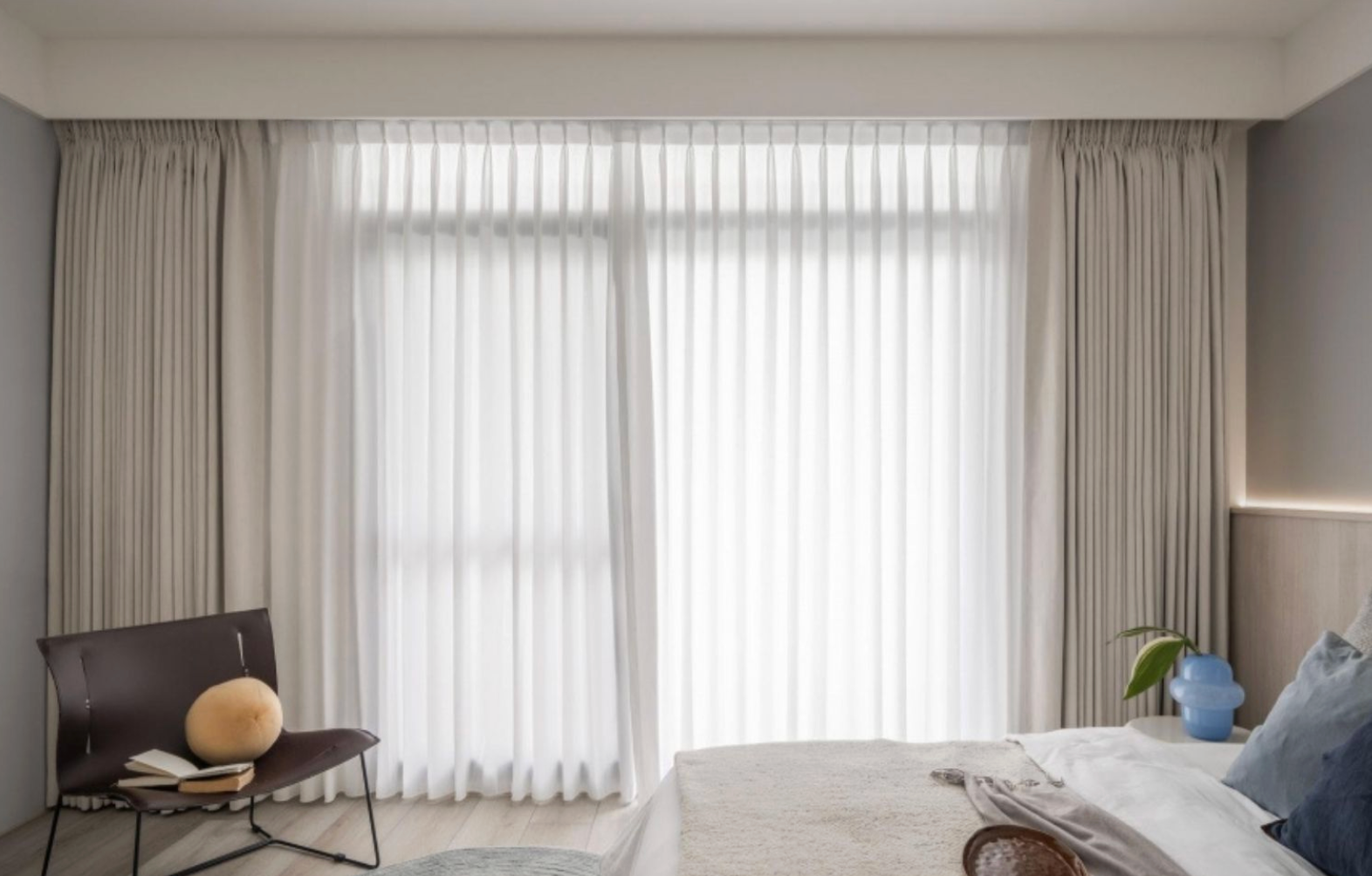臥室近景,雙層窗簾內層為白色柔紗簾,外層為淺灰色落地布簾,整體呈現垂直波浪褶皺;左側有皮質單椅,床邊放著藍色玻璃花瓶。
